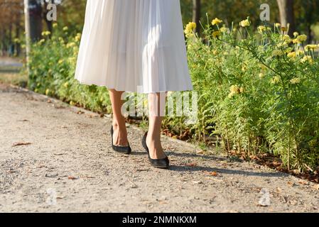 Femme dans une jupe plissée blanche et des stilettos en daim noir se précipitent à la maison après le travail à travers un parc le long d'une allée de fleurs jaunes, section basse. Banque D'Images