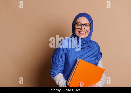 Positive mature enseignante dans le hijab bleu, souriant un sourire crasseux regardant la caméra, tenant des livres, fond beige Banque D'Images