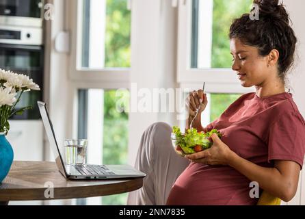 Jolie jeune femme mangeant de la salade à table dans la salle à manger avec un ordinateur portable devant elle. Un mode de vie sain pendant la période de maternité Banque D'Images