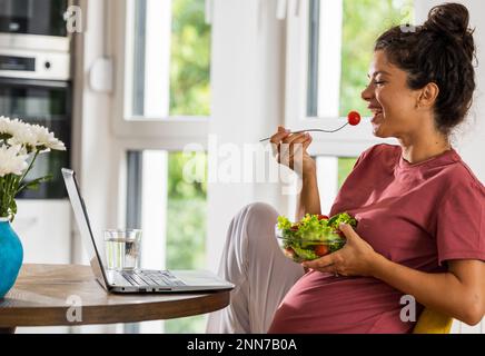 Jolie jeune femme mangeant de la salade à table dans la salle à manger avec un ordinateur portable devant elle. Un mode de vie sain pendant la période de maternité Banque D'Images