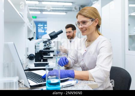Femme scientifique mature en couche blanche au travail, assistante de laboratoire utilisant un microscope et un ordinateur portable au travail, scientifique travaillant à l'intérieur de la recherche de laboratoire. Banque D'Images
