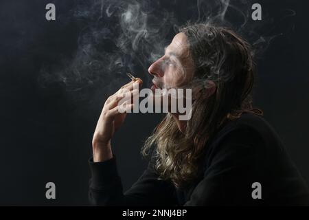 Studio portrait sur fond noir de l'homme d'âge moyen avec de longs cheveux fume la cigarette Banque D'Images