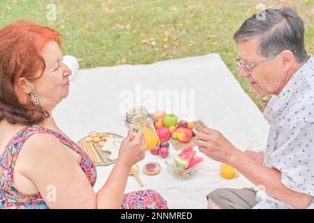 Un heureux couple latin senior ayant un petit déjeuner pique-nique sain avec du pain de grain entier, du jus d'orange et des fruits variés. Banque D'Images