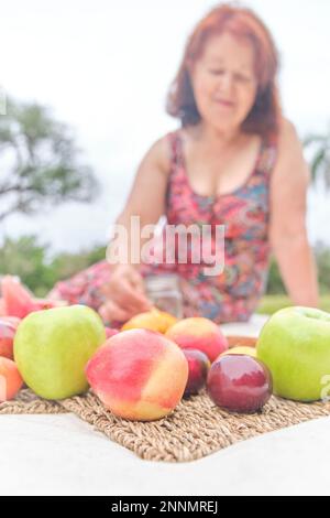 Femme mature ayant un petit déjeuner pique-nique avec une variété de fruits frais, pommes, pêches, mandarines, prunes, pastèque. Concentrez-vous sur eux. Concepts : santé Banque D'Images
