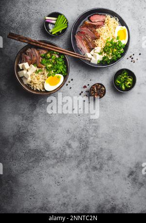 Deux bols de savoureuses nouilles asiatiques soupe ramen avec bouillon, tofu, porc, oeuf sur fond gris rustique en béton, espace pour le texte, gros plan, vue de dessus. Chaud Banque D'Images