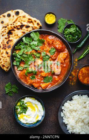 Plat indien traditionnel poulet tikka masala avec viande épicée au curry dans un bol, riz basmati, pain naan, sauce au yaourt raita sur fond rustique sombre Banque D'Images