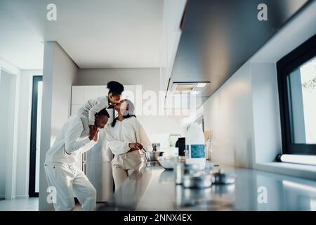 Détente en famille au petit déjeuner. Maman embrasse sa fille des épaules de papa. Famille ayant des moments ludiques pendant que maman prépare le petit déjeuner le matin. Banque D'Images