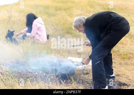 L'ancien homme de cheveux blanc tourne le gril sur la boîte à côté de la fille avec le chien dans l'herbe de champ. Banque D'Images