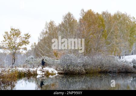 Le pêcheur à la ligne moderne dans un costume de protection bleu se dresse sur les rives enneigées des birches de montagne dans la neige au bord de l'eau du lac pour tourner la Russie Banque D'Images