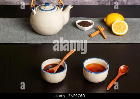 Deux tasses de thé, une verte et une rouge au premier plan, en arrière-plan une théière avec des citrons, des morceaux de cannelle et du thé moulu. Banque D'Images