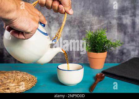 Mains tenant une théière et versant du thé dans une tasse, sur une table en bois bleu. Banque D'Images