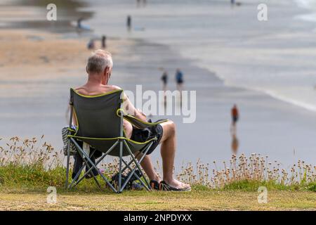 Un ancien homme de vacances assis dans une chaise pliante sur la côte surplombant la plage de Fistral à Newquay, en Cornouailles, au Royaume-Uni. Banque D'Images