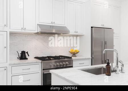 Un détail de cuisine avec des armoires blanches, une cuisinière et une hotte en acier inoxydable, des comptoirs en marbre et un dosseret, et un évier chromé. Banque D'Images