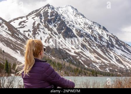 Femme assise dans la région sauvage du nord du Canada avec des sommets enneigés admirant en arrière-plan et le paysage sec du printemps Banque D'Images