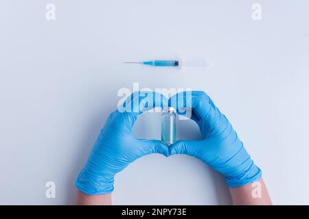 Sur fond blanc isolé, les mains des médecins en gants médicaux bleus représentent le geste de la forme du cœur. Le médecin tient une ampoule avec le vaccin contre v Banque D'Images