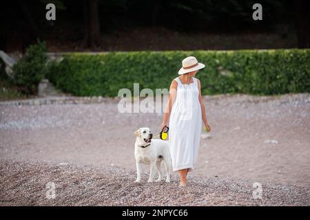 Future mère enceinte marchant le long du sable de la plage près de la mer avec Golden Retriever. Femme millénaire en chapeau blanc tient le chien sur la laisse. Conce Banque D'Images