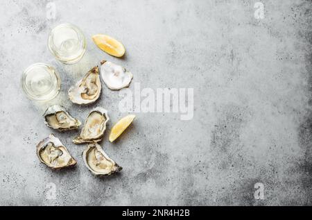 Gros plan d'une demi-douzaine d'huîtres fraîches ouvertes et de coquillages avec des quartiers de citron, deux verres de vin blanc ou de champagne, vue de dessus, béton rustique gris Banque D'Images