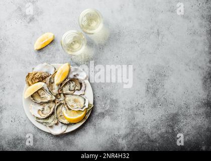 Gros plan d'une demi-douzaine d'huîtres fraîches ouvertes et de coquillages avec des quartiers de citron sur une assiette, deux verres de vin blanc, vue de dessus, béton rustique gris Banque D'Images