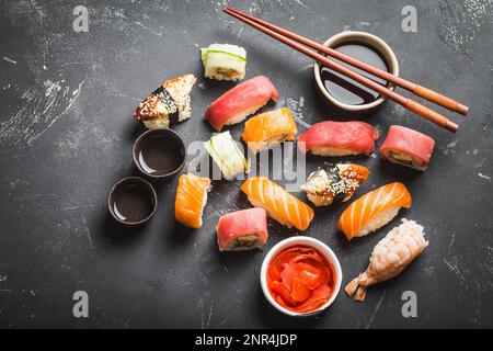 Vue de dessus d'un assortiment de sushis japonais avec petits pains, nigiri, sauce soja, gingembre, baguettes, deux tasses de saké traditionnel sur béton noir Banque D'Images