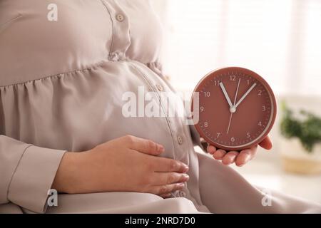 Jeune femme enceinte tenant l'horloge près de son ventre à la maison, en gros plan. Il est temps de donner naissance Banque D'Images