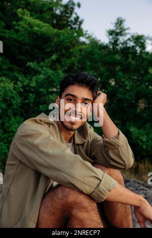 Portrait d'un jeune homme souriant se penchant la tête sur le coude tout en étant assis contre les plantes Banque D'Images