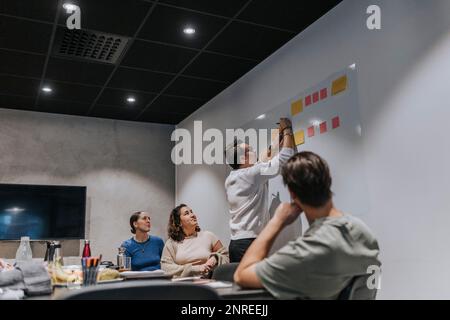 Un jeune homme d'affaires écrit sur une note adhésive de collègues hommes et femmes dans la salle de bord Banque D'Images