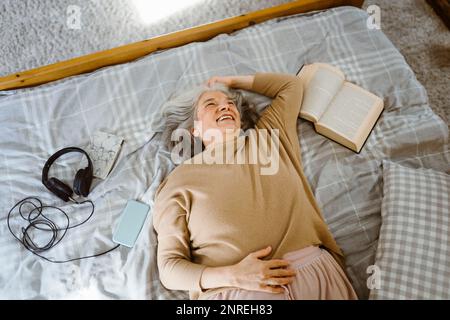 Bonne femme âgée avec livre et écouteurs couchés sur le lit Banque D'Images