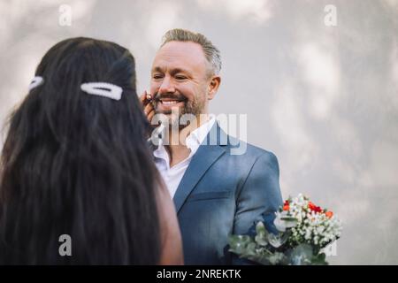 Vue arrière de la mariée touchant la joue souriante du marié contre le mur le jour du mariage Banque D'Images