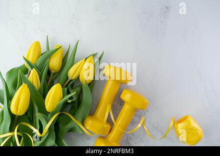 Des haltères de gym et des fleurs de tulipes jaunes en cadeau pour la fête des femmes ou l'anniversaire. Entraînement physique sain, entraînement sportif plat avec espace de copie. Banque D'Images