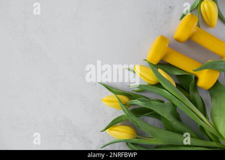 Des haltères de gym et des fleurs de tulipes jaunes en cadeau pour la fête des femmes ou l'anniversaire. Entraînement physique sain, entraînement sportif plat avec espace de copie. Banque D'Images