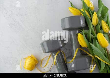 Des haltères de gym et des fleurs de tulipes jaunes en cadeau pour la fête des femmes ou l'anniversaire. Entraînement physique sain, entraînement sportif plat avec espace de copie. Banque D'Images