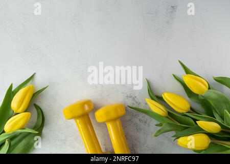 Des haltères de gym et des fleurs de tulipes jaunes en cadeau pour la fête des femmes ou l'anniversaire. Entraînement physique sain, entraînement sportif plat avec espace de copie. Banque D'Images