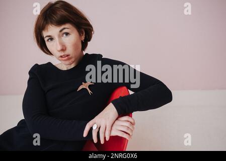 une femme vêtue d'une robe noire est assise sur une chaise, sur fond rose Banque D'Images