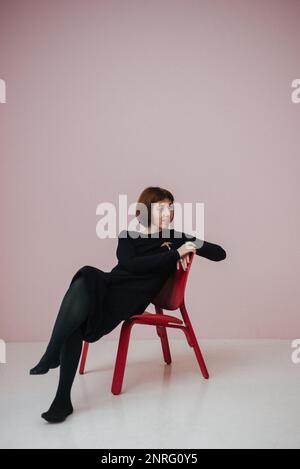 une femme vêtue d'une robe noire est assise sur une chaise, sur fond rose Banque D'Images