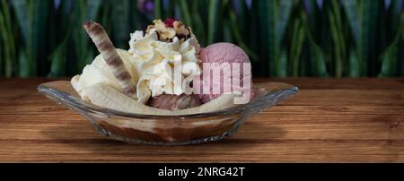 Fente de banane garnie de crème fouettée et sorbet au chocolat en canard de verre transparent sur table en bois avec des plantes vertes défocused de fond. Image avec Banque D'Images