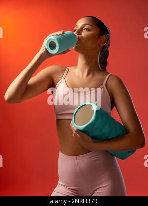 Étancher sa soif après une bonne session. Photo en studio d'une jeune femme sportive buvant de l'eau et tenant un rouleau en mousse sur fond rouge. Banque D'Images