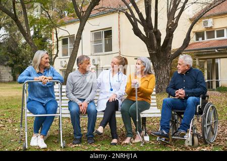 Médecin parlant à un groupe heureux de personnes âgées assis sur un banc dans le jardin de la maison de soins infirmiers Banque D'Images