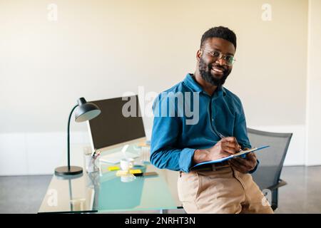 Concept d'entrepreneuriat réussi. Homme d'affaires noir enthousiaste tenant une presse-papiers avec des documents et souriant, espace de copie Banque D'Images