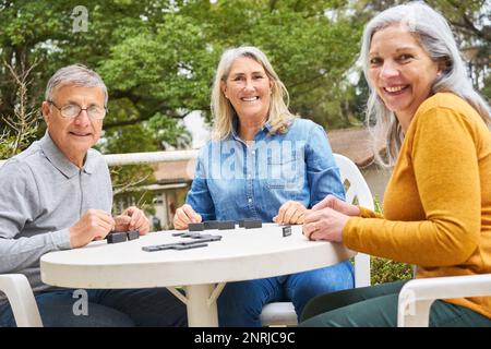 Portrait d'amis aînés heureux jouant des dominos sur la table dans le jardin de la maison de soins infirmiers Banque D'Images