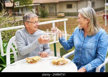 Homme et femme joyeux et senior toaster un verre d'eau tout en prenant un repas dans le jardin de la maison de retraite Banque D'Images