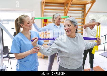 Femme thérapeute souriante aidant une femme âgée faisant de l'exercice avec un groupe de résistance au centre de réadaptation Banque D'Images