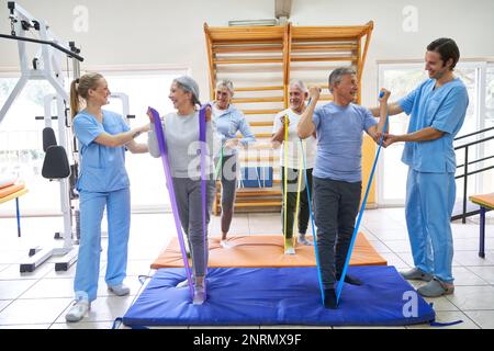 Des physiothérapeutes souriants aident les personnes âgées à faire de l'exercice avec des bandes élastiques au centre de réadaptation Banque D'Images