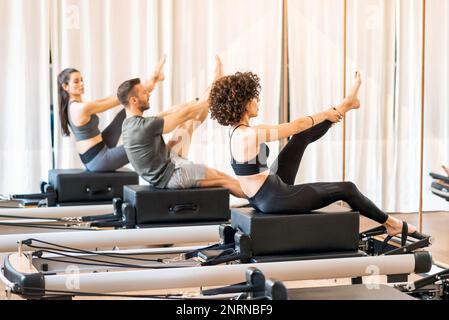 Des personnes sportives concentrées dans les oreilles actives qui étirent les jambes sur les pillates reformer pendant la classe de groupe Banque D'Images