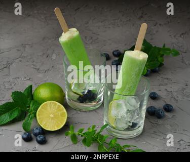 Kiwi, lime, glace à la menthe ou popsicles faits maison avec des bleuets mûrs sur une table en verre et en ciment de béton gris ou à l'arrière. Nourriture saine d'été conc Banque D'Images