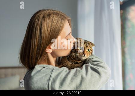 Décontracté jeune femme Blonde tient l'épaule un mignon chat écossais de Tabby à l'oeil vert et l'épouse à la maison à la fenêtre, le concept de l'amour et de prendre soin des animaux de compagnie Banque D'Images