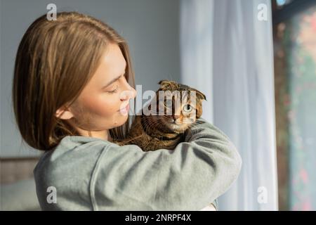 Décontracté jeune femme Blonde tient l'épaule un mignon chat écossais de Tabby à l'oeil vert et l'épouse à la maison à la fenêtre, le concept de l'amour et de prendre soin des animaux de compagnie Banque D'Images