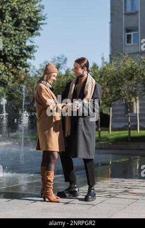 pleine longueur de bonne blonde femme dans le manteau et le chapeau tenant les mains avec le boyfriend tatoué dehors, image de stock Banque D'Images