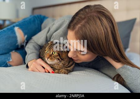 Happy Young blonde femme couché dans son lit dans un chandail un chien de train domestique mignon jouant écossais tabby chat à la maison, concept aimant et les animaux de compagnie Banque D'Images