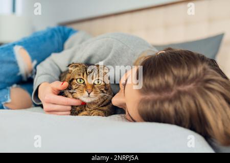 Happy Young blonde femme couché dans son lit dans un chandail un chien de train domestique mignon jouant écossais tabby chat à la maison, concept aimant et les animaux de compagnie Banque D'Images