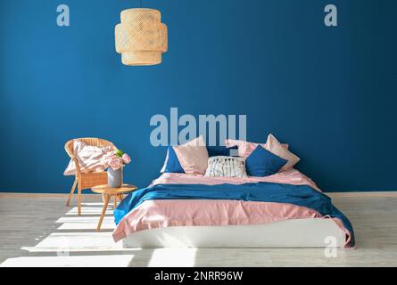 Belle chambre moderne. Grand lit double, mur bleu foncé, couvre-lit rose, couverture bleue, oreillers, vase décoratif, fleurs sur une petite table en bois, passé en osier Banque D'Images